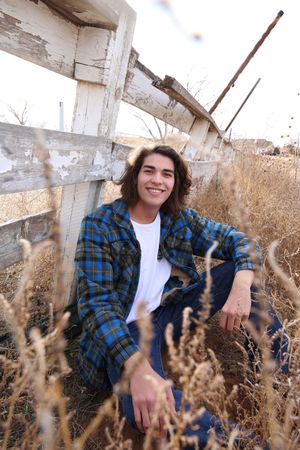 Smiling young man with dark wavy hair and a blue plaid flannel sits in dry golden grass beside a weathered white wooden fence.