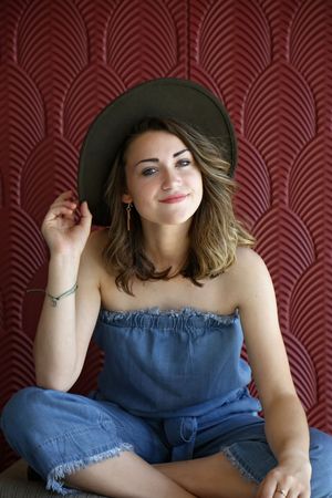 Smiling woman in a wide-brim hat and denim strapless romper sits in front of a red art deco patterned wall.