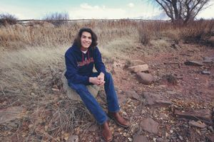Smiling young man with dark hair, navy hoodie and jeans sits on a rock in a dry desert field with distant trees.