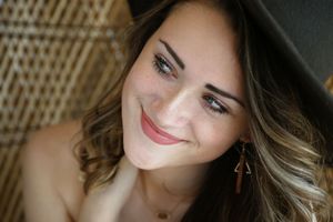 Close-up portrait of a smiling woman with wavy highlighted hair and drop earrings sitting near a woven wicker chair.