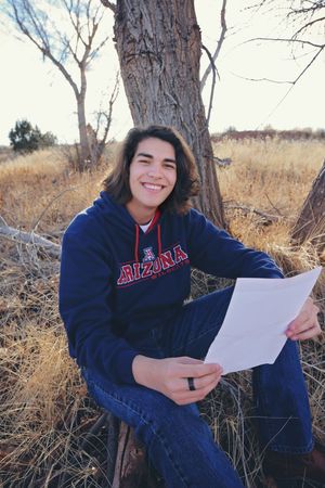 Smiling young man in an Arizona sweatshirt sits against a tree in a dry grassy field holding a sheet of paper.