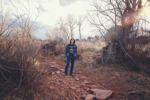 Young man in a denim jacket and jeans stands on a rocky dirt path surrounded by bare trees and dry brush at sunset.