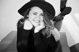 Black and white portrait of a smiling woman in a black hat and sweater lying on her stomach with her chin on her fists.