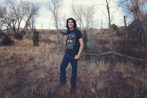 Young man in a graphic black t-shirt and blue jeans stands in a dry grassy field with bare trees and desert shrubs.