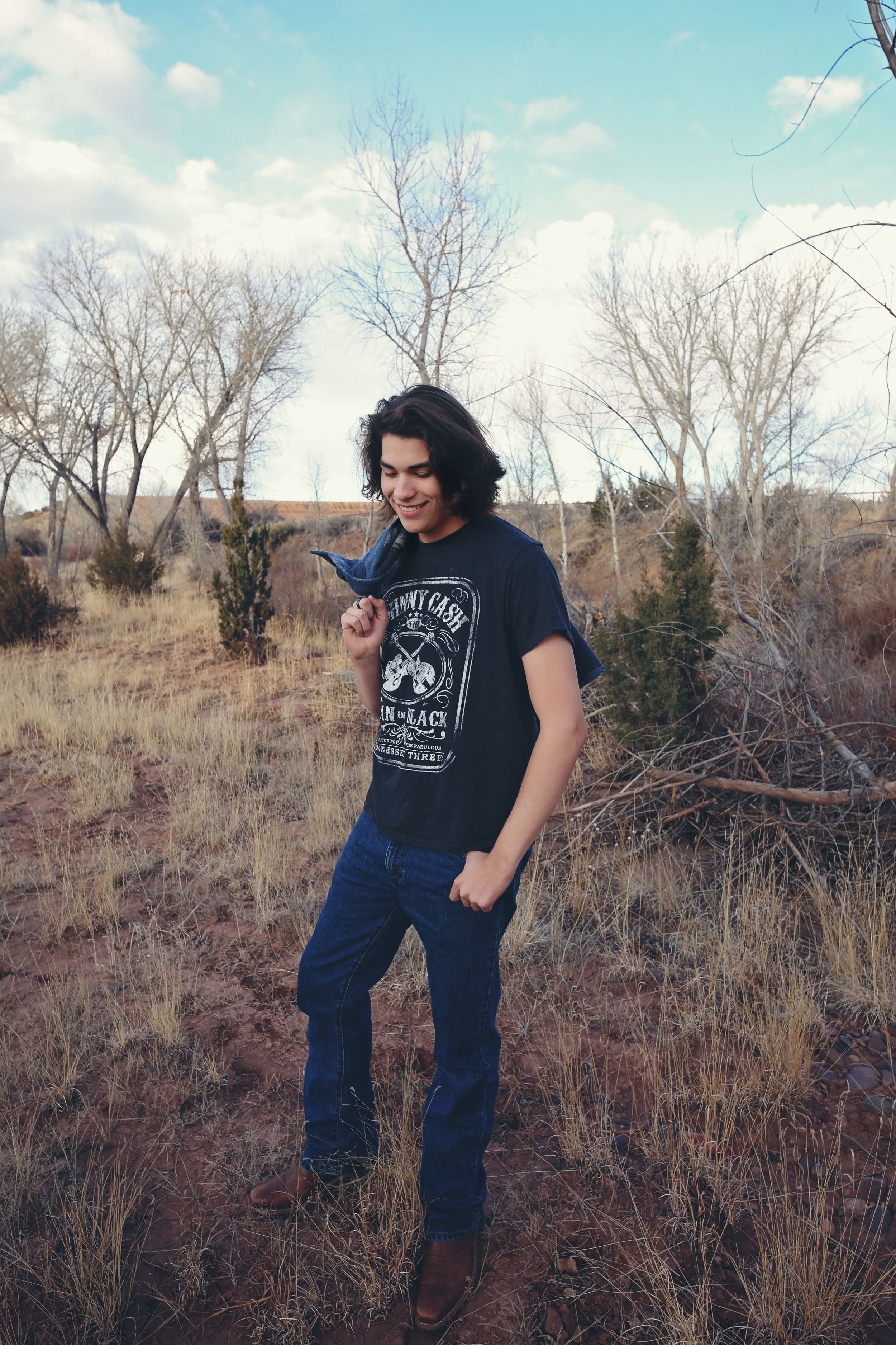 Young man with dark hair in a graphic t-shirt and jeans standing in a dry desert field with bare trees and a blue sky.