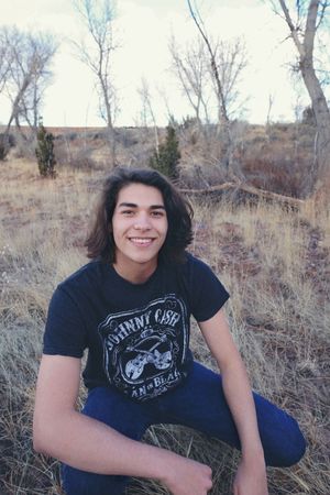 Smiling young man with shoulder-length dark hair and a Johnny Cash t-shirt crouches in a dry grassy desert landscape.