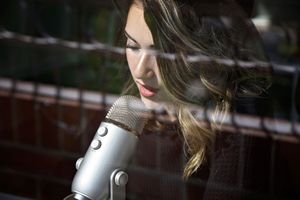 Woman with long wavy hair sings into a silver studio microphone behind a glass window with soft reflections.