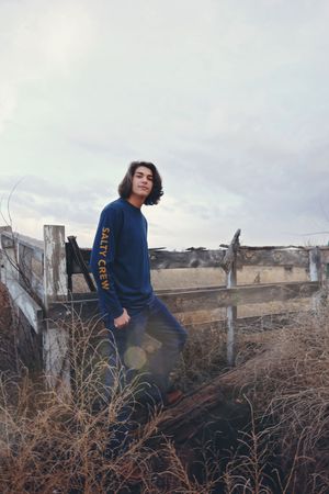 Young man with shoulder-length dark hair leans on a weathered wooden fence in a dry grass field under a cloudy sky.