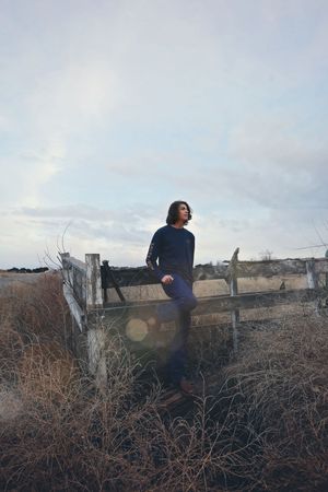 Young man in a navy sweatshirt sits on a rustic wooden fence surrounded by tall dry grass beneath a soft, cloudy sky.