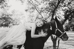 Black and white photo of a woman in a flowing gown lying across the back of a horse in a tree-filled pasture.