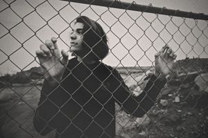 Black and white portrait of a young man with dark hair gripping a chain-link fence with a rugged landscape behind him.