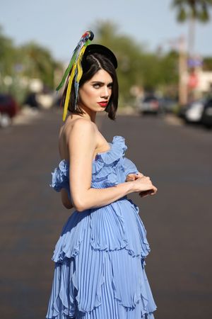 Woman in a blue ruffled gown and a feathered parrot headpiece poses on a sunny street with palm trees in the background.