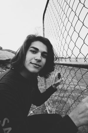 Black and white portrait of a smiling young man with wavy hair touching a chain-link fence beside the water.