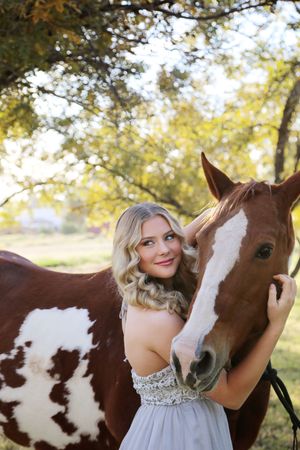 Smiling blonde woman in a gray gown embraces a chestnut and white paint horse under golden autumn trees.