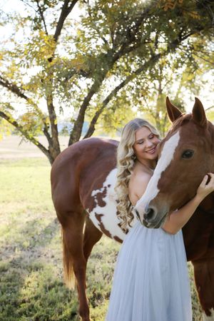 Blonde woman in a gray dress smiles as she hugs a chestnut paint horse beneath a leafy tree in a grassy field.