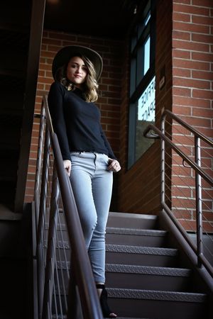 A young woman in a wide-brim hat, black top and gray jeans poses on a metal staircase beside a red brick building.