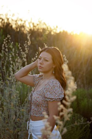 Young woman in a floral crop top and white skirt poses with her hand in her hair among tall wild grasses at golden hour sunset.