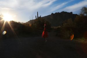 A dancer in a red dress stands on a desert trail at sunrise with rugged mountains and glowing sun flare in the background.