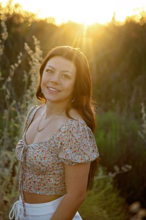 Smiling young woman in a floral crop top and necklace is backlit by warm golden sunlight streaming through a wild grass field.