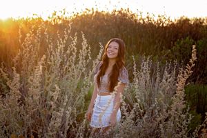 Young woman in a floral top and white skirt smiles while standing among tall silvery grasses glowing in warm sunset light.