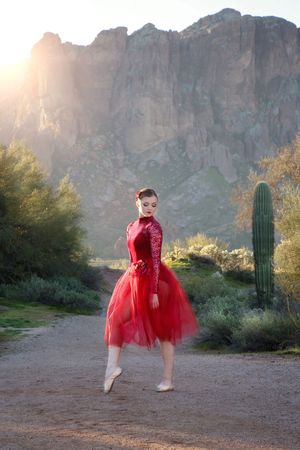 A ballerina in a red lace dress and tulle skirt poses on pointe on a desert path with a saguaro cactus and sunlit cliffs behind.