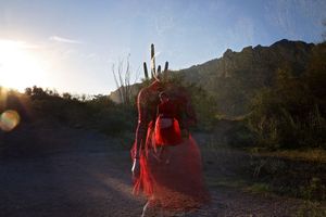 A double-exposure portrait of a dancer in red on a desert trail at sunset, with mountains and a cactus in the background.