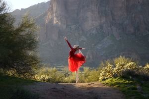 A dancer in a flowing red dress leaps with outstretched arms on a desert trail beneath towering rocky cliffs.