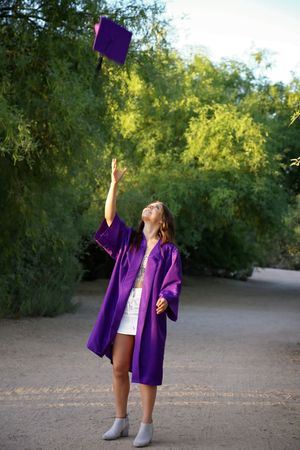 Graduate in a purple gown tosses her graduation cap into the air on a tree-lined dirt path for a joyful senior portrait.