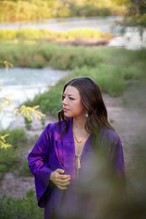 A young woman in a purple satin robe stands among green riverside plants, gazing to the side in soft natural light.