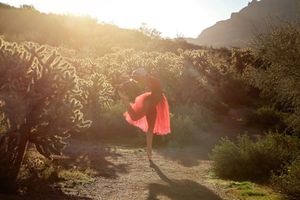 A dancer in a flowing red skirt poses mid-step on a sunlit desert trail lined with glowing cholla cacti.