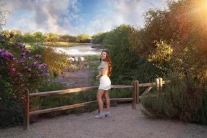 Smiling young woman in a white skirt stands by a wooden fence near a pond with colorful flowers under a dramatic sunset sky.