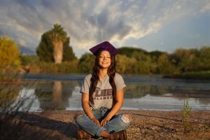 A smiling graduate in a purple cap sits cross-legged on a lakeshore at sunset with trees and reflections in the water.