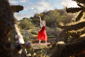 Ballerina in a flowing red dress dances with one arm raised in a sunlit desert landscape framed by cholla cactus.