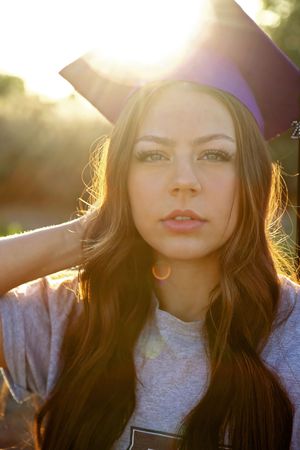 Sun-flared portrait of a young woman with long hair wearing a purple graduation cap and a gray shirt outdoors.