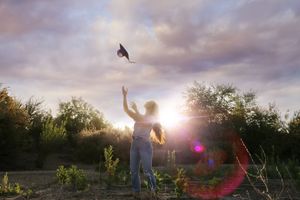 Young woman tosses her graduation cap into the air at sunset with warm lens flare, trees and a pastel cloudy sky behind.