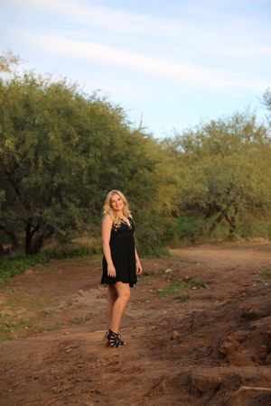 Smiling blonde woman in a short black dress and strappy sandals stands on a dirt path beside green desert trees.