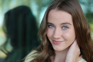Close-up portrait of a young woman with blue eyes and wavy light brown hair smiling gently while touching her neck.