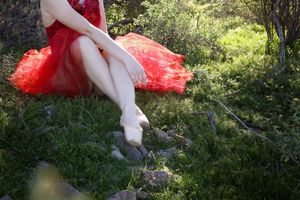 Woman in a red tulle dress sits on a grassy hillside with her legs crossed and hands resting on her ankle.