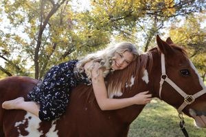 Smiling young woman with long wavy blonde hair lies across the back of a brown and white paint horse under autumn trees.