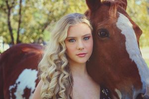 Young woman with long curly blonde hair poses closely beside a brown and white horse in a sunlit outdoor portrait.