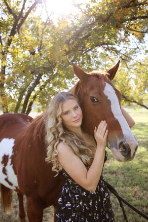 Young woman with curly blonde hair in a floral dress rests her hand on a chestnut paint horse in golden sunlight.