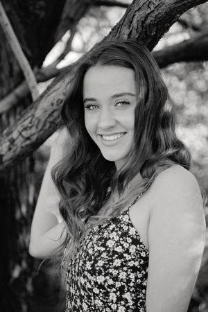 Black and white portrait of a smiling young woman with long wavy hair in a floral top posing beside a large tree trunk.
