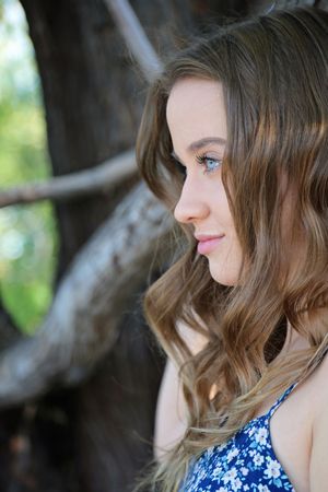 Side profile portrait of a young woman with wavy brown hair in a blue floral top gazing away beside a gnarled tree.