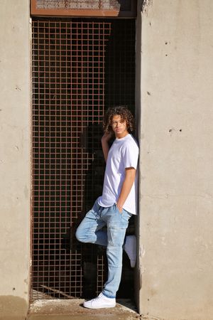 Young man with curly hair in a white t-shirt and light jeans leans against a concrete wall beside a metal grate.