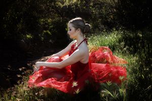Ballerina in a flowing red tulle dress sits on sunlit green grass, her head bowed, against a dark leafy background.