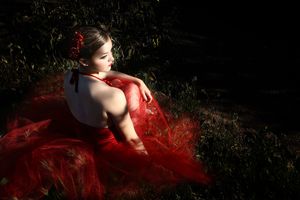 Woman in a flowing red tulle dress with a red flower in her hair sits on dark grass in dramatic evening light.