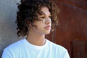 Young man with curly dark hair in a white t-shirt gazes thoughtfully to the side beside a rusted metal wall.