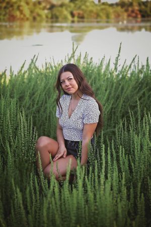 Young woman in a white patterned blouse sits among lush green plants beside a calm lake during a senior portrait session.