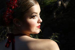 Close-up portrait of a woman with red lipstick and red floral hair accessory looking over her bare shoulder in dramatic light.