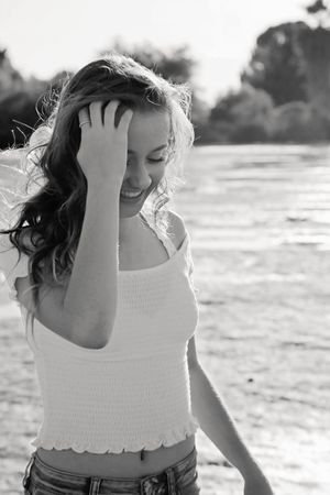 Black and white photo of a laughing young woman in a smocked white top and jeans brushing her hair back by a sunlit lake.
